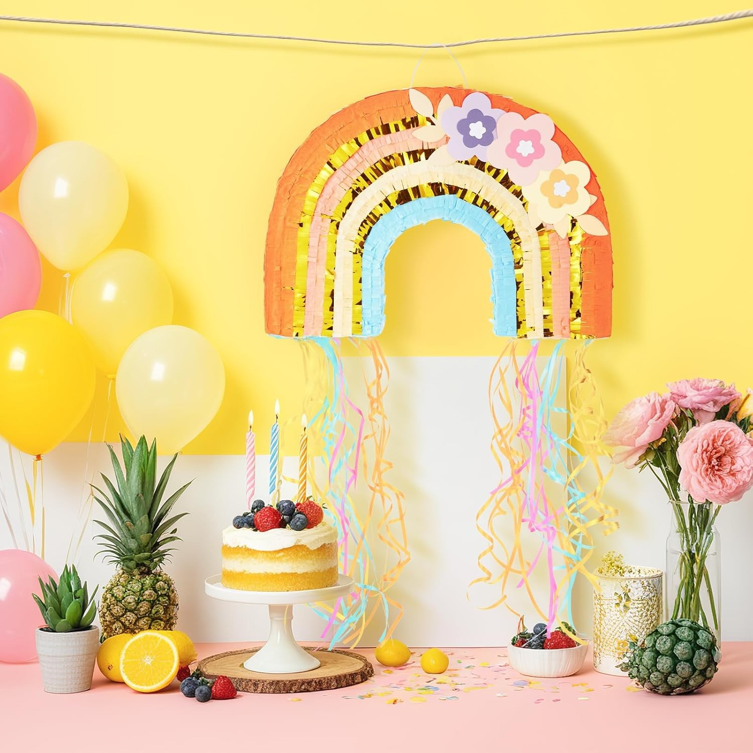 Birthday setup with cake, balloons, and rainbow pinãta against a yellow wall.